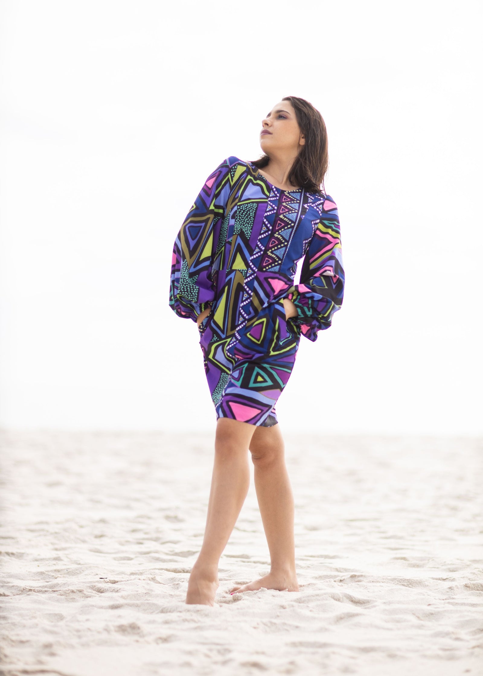 Original Print Shift Dress with Japanese Silk featuring a vibrant geometric print in shades of purple, blue, pink, green, and black. This relaxed shift dress is crafted from luxurious Japanese silk with voluminous long sleeves and a flattering above-the-knee silhouette. Styled barefoot on a sandy beach, the look captures effortless elegance, artistic craftsmanship, and resort-inspired sophistication.