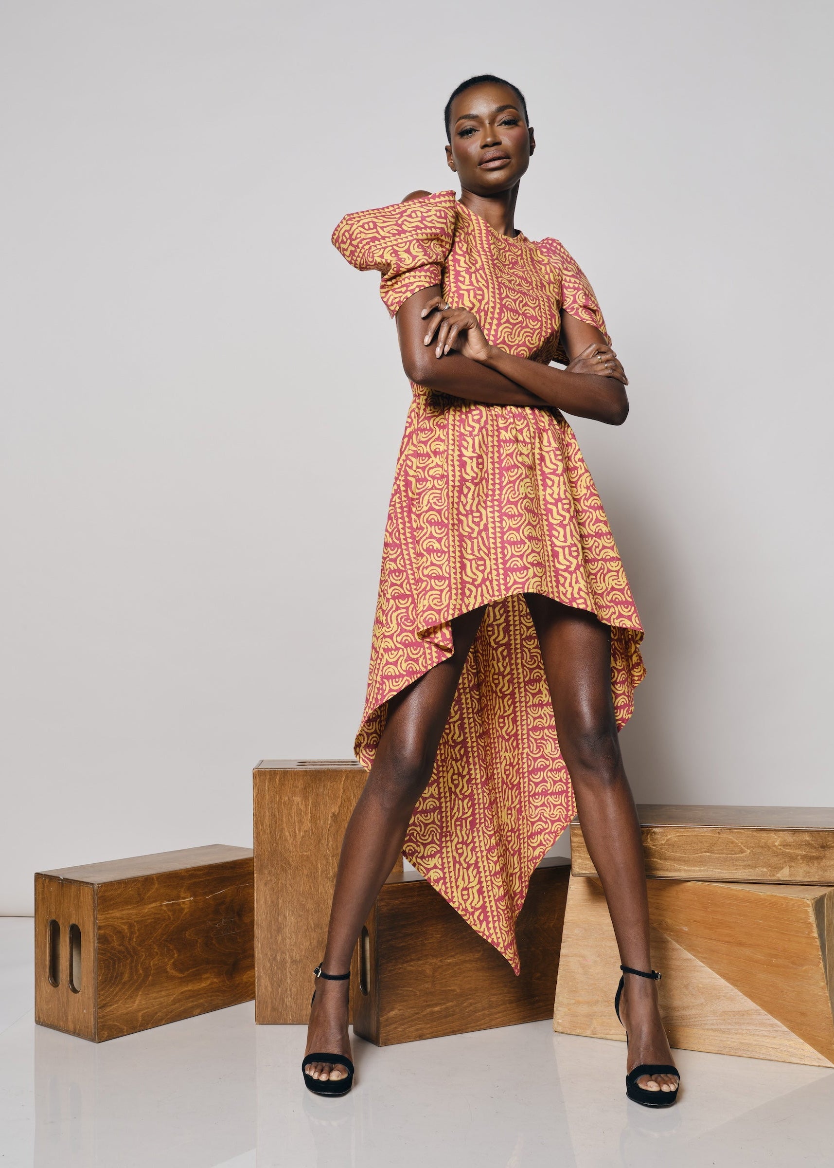 Black model wearing the KAHINDO Simien Dress, a pink and yellow African-inspired print high-low dress with dramatic puff sleeves, styled with black heels in a minimalist studio setting.