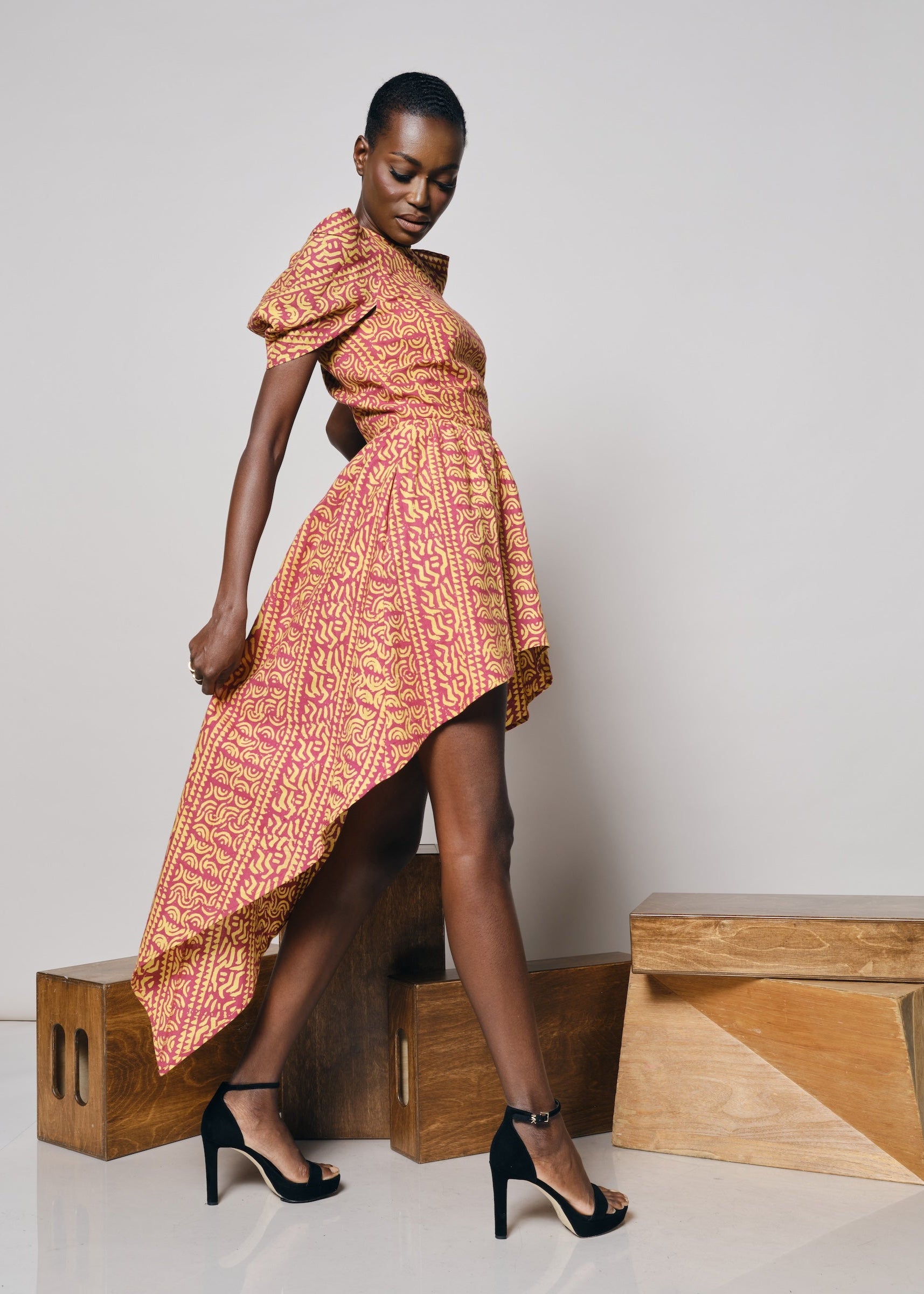 Black model wearing the KAHINDO Simien Dress, a pink and yellow African-inspired print high-low dress with dramatic puff sleeves, styled with black heels in a minimalist studio setting.