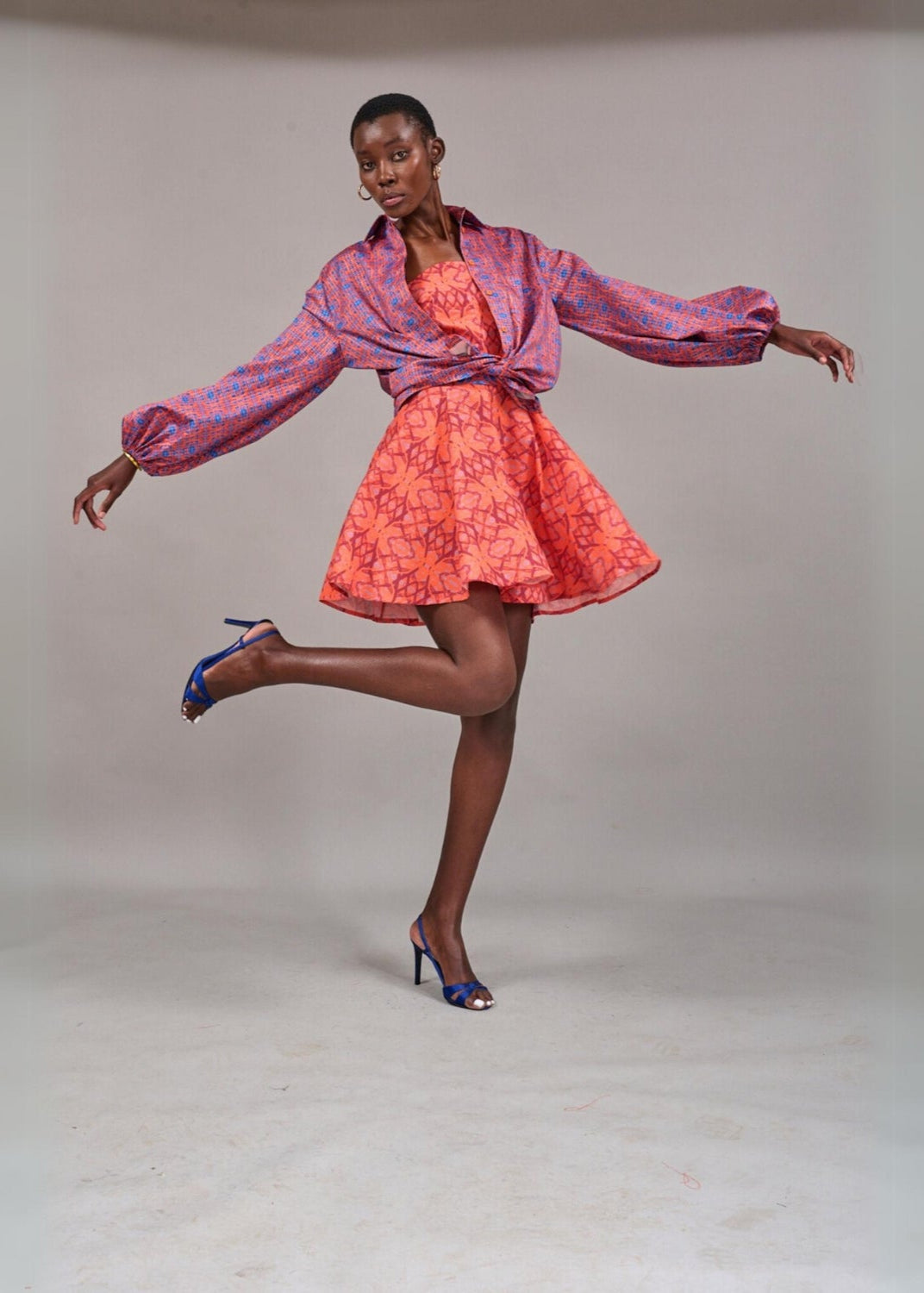 Model wearing the KAHINDO Stellenbosch Button Down, a lightweight long-sleeve printed button-down shirt with a purple and blue geometric pattern, styled open over a coral dress in a minimalist studio.