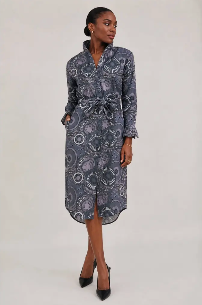 Zambia Shirtdress in a gray geometric African-inspired print featuring a collared button-front design, long sleeves, self-tie waist belt, midi length, and side pockets, modeled with black pointed-toe heels against a neutral studio background.