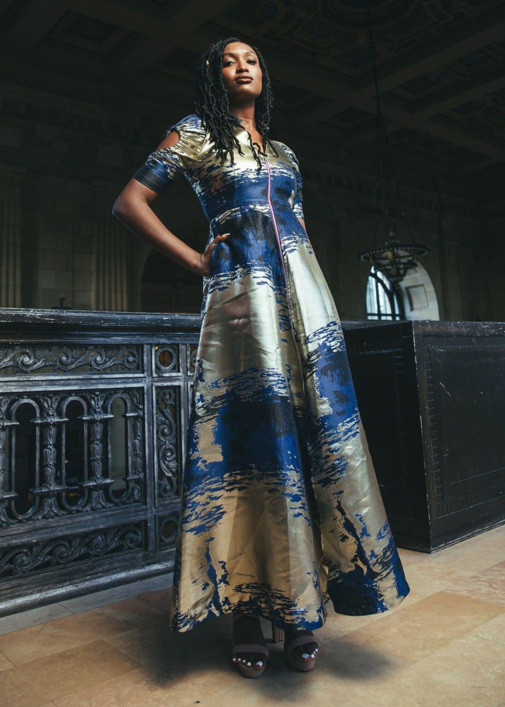 Model showcasing the Cleopatra Maxi Dress with blue and gold patterns in an indoor setting