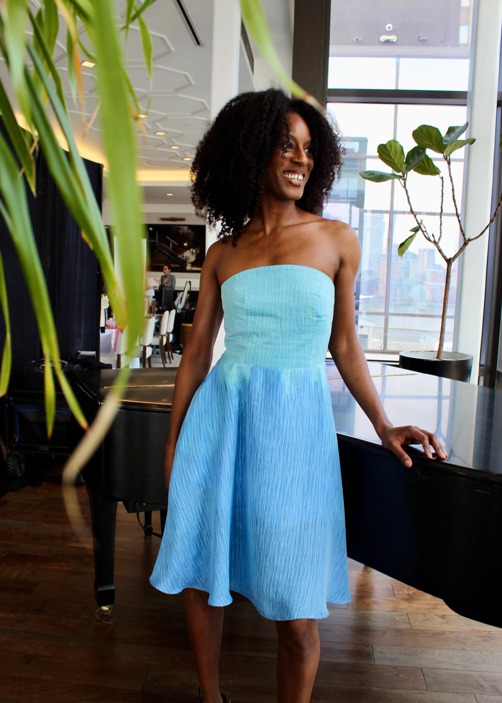 Woman in a light blue dress standing indoors with plants and a piano in the background