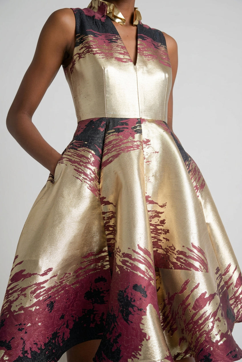 Woman wearing a sleeveless dress with gold, pink, and black abstract pattern against a white curtain background.