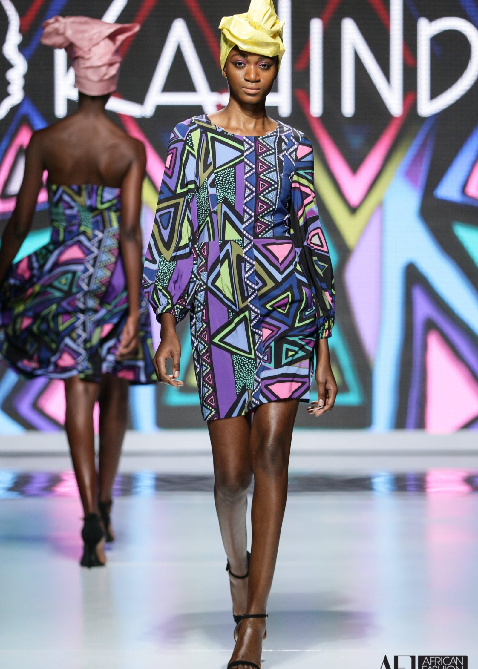 Original Print Shift Dress with Japanese Silk featuring a vibrant geometric print in shades of purple, blue, pink, green, and black. This relaxed shift dress is crafted from luxurious Japanese silk with voluminous long sleeves and a flattering above-the-knee silhouette. Styled barefoot on a sandy beach, the look captures effortless elegance, artistic craftsmanship, and resort-inspired sophistication.