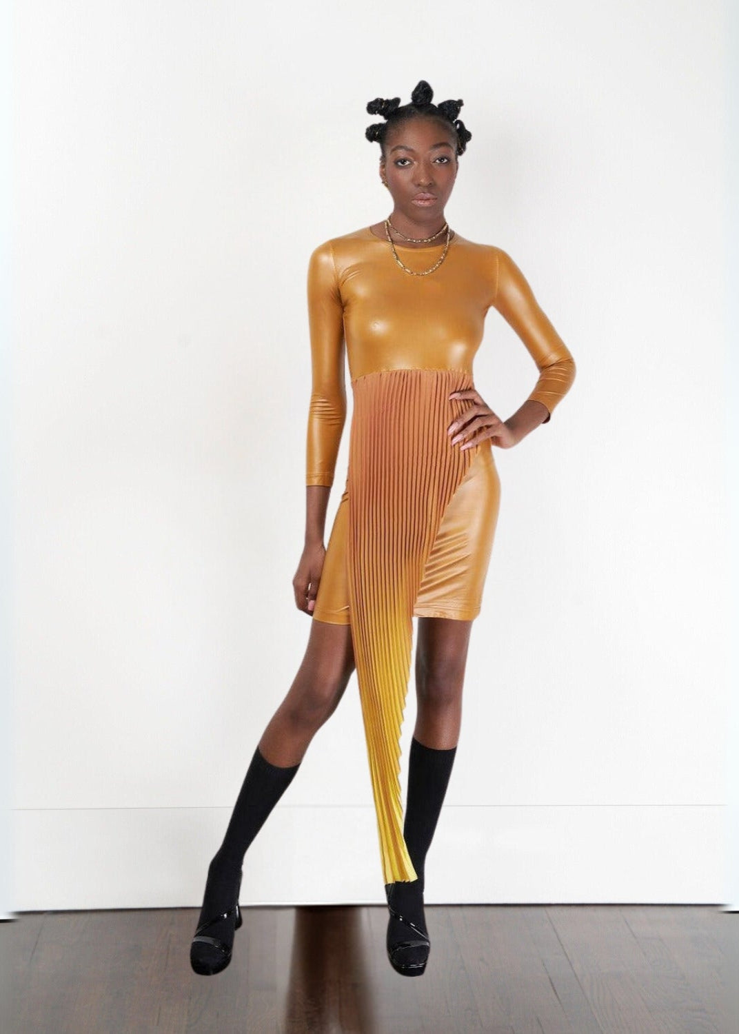 Sibayi Midi Dress in a metallic gold stretch fabric featuring a fitted long-sleeve silhouette with a unique asymmetrical pleated panel cascading from the waist. Styled with black knee-high socks, black platform shoes, and layered gold necklaces against a clean white studio background, showcasing the dress's sculptural, contemporary design.