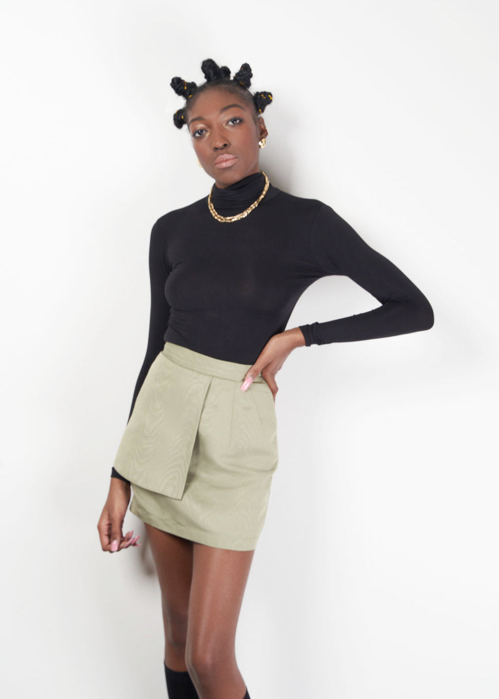 Model wearing the KAHINDO Tana Skirt, a sage green moiré taffeta mini skirt with a structured wrap-front silhouette and invisible back zipper, styled with a black turtleneck in a clean studio setting.