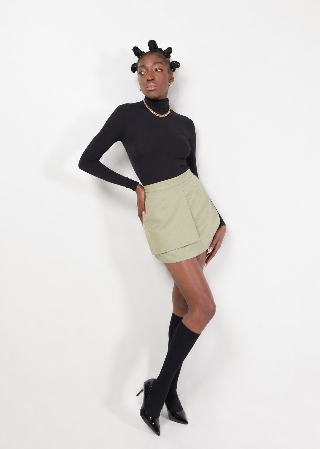 Model wearing the KAHINDO Tana Skirt, a sage green moiré taffeta mini skirt with a structured wrap-front silhouette and invisible back zipper, styled with a black turtleneck in a clean studio setting.